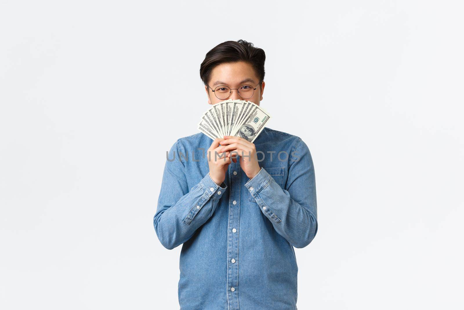 Smiling satisfied asian guy in glasses, hiding face behind money and peeking at camera cunning, earn cash for working freelance, making investment, close deposit, standing white background by Benzoix