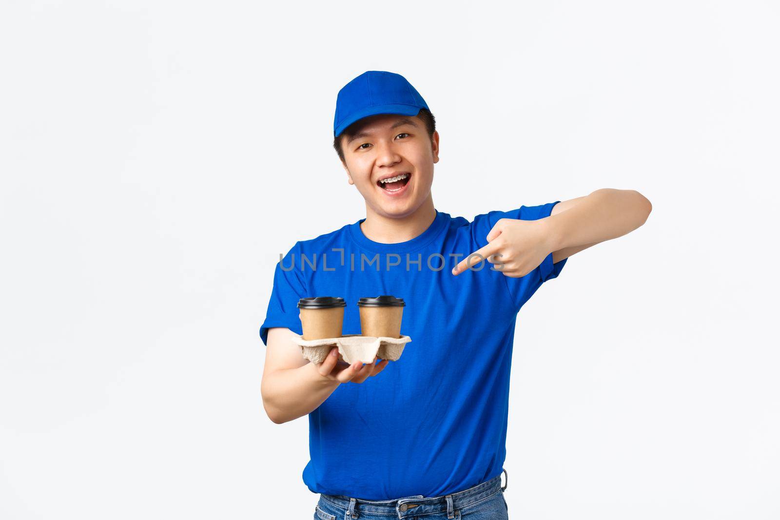 Online orders, takeaway and employees concept. Friendly cheerful male courier in blue uniform ensure fast delivery from restaurants or cafes, pointing finger at coffee and smiling, white background.