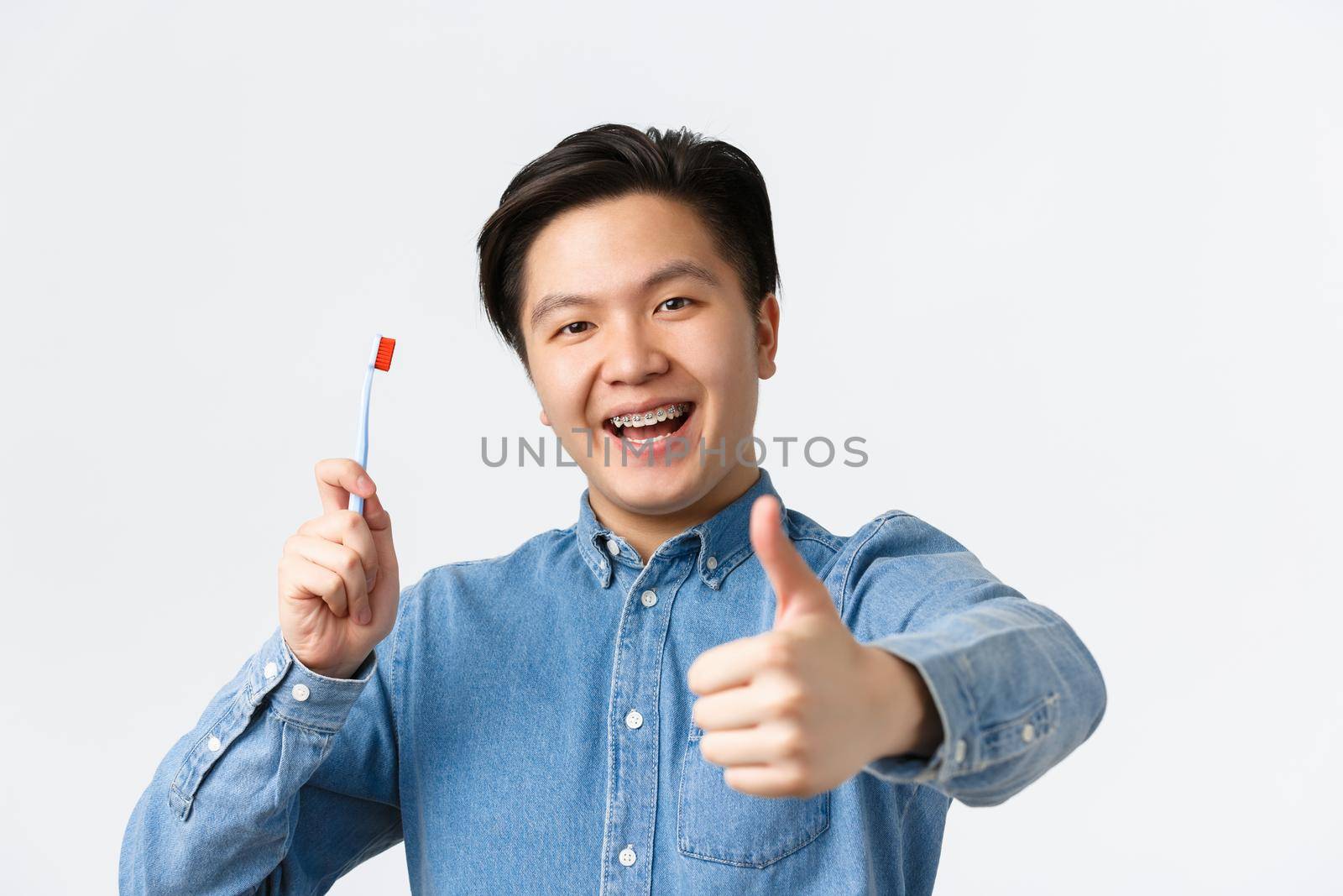 Orthodontics, dental care and hygiene concept. Close-up of satisfied happy asian guy holding toothbrush, smiling with teeth braces and showing thumbs-up in approval, white background by Benzoix