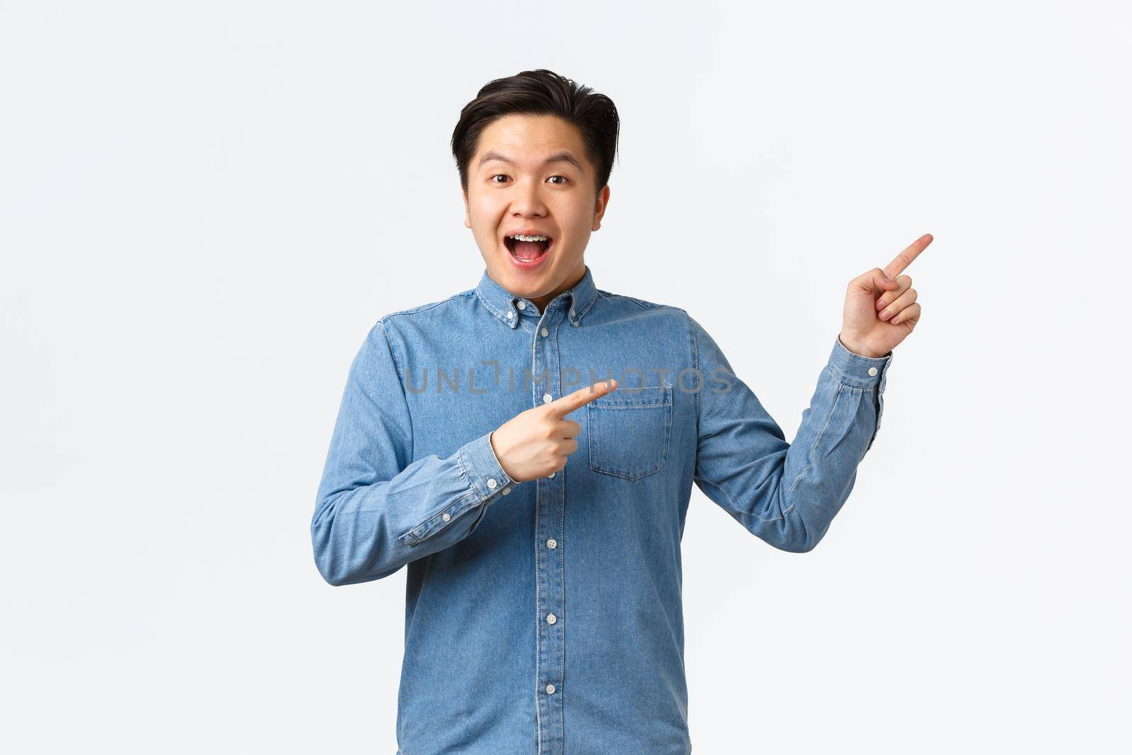 Excited and happy asian man making big announcement. Guy with braces smiling amused and pointing fingers upper right corner. Male student showing courses or online link, white background.