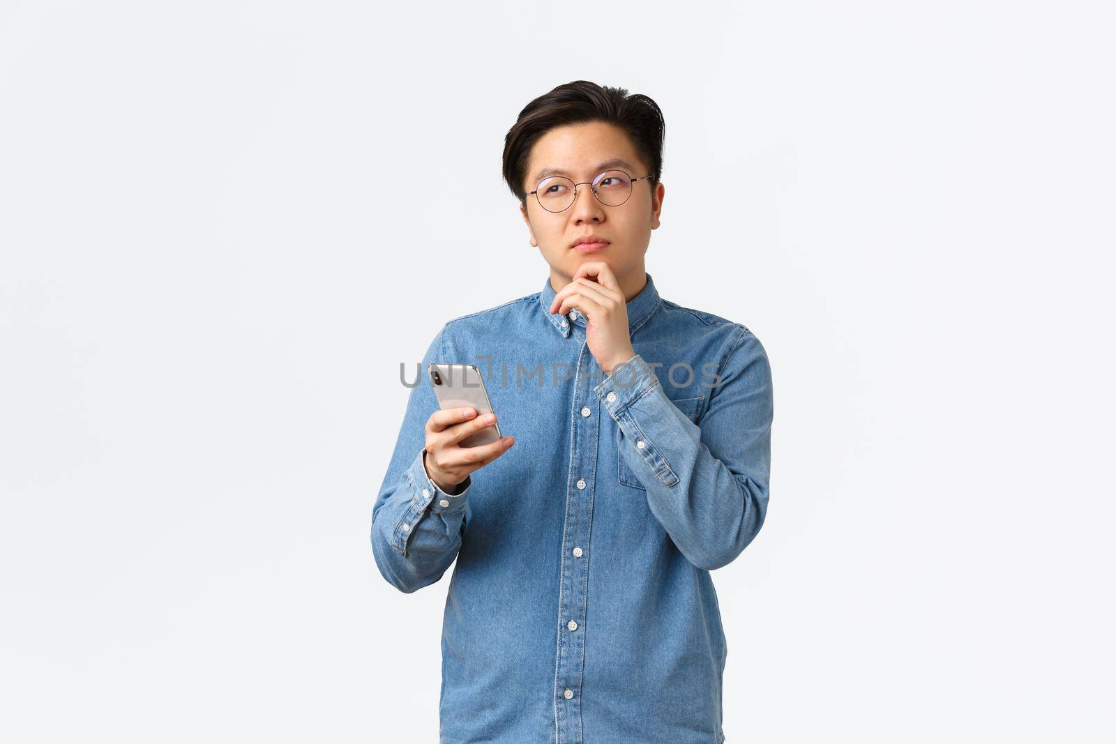 Thoughtful creative asian man in glasses thinking while making post on social media, looking away, pondering or making decision, holding smartphone, choosing something in internet by Benzoix