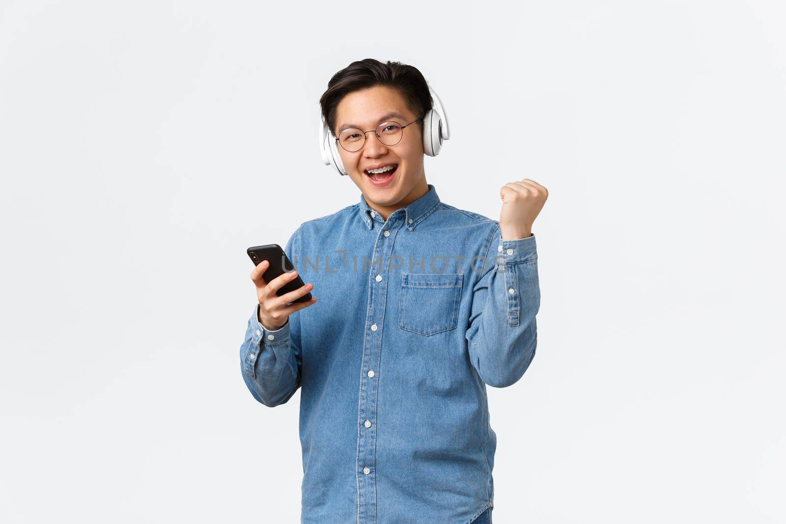 Lifestyle, leisure and technology concept. Winning satisfied asian man in glasses, listening music in wireless headphones, using mobile phone, smiling and showing fist pump triumph gesture at camera.