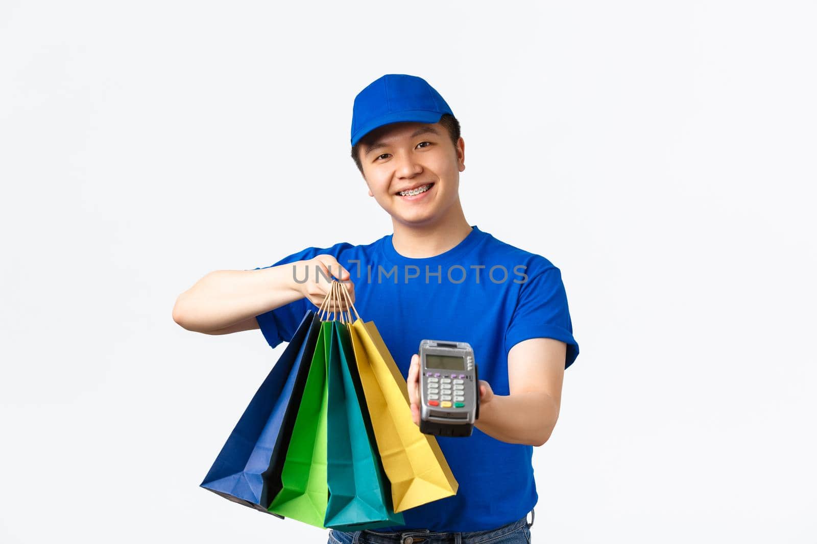 Contactless payment, shopping and home delivery concept. Smiling pleasant asian courier in blue uniform bring client order to doorstep, handing packages and POS terminal for paying.