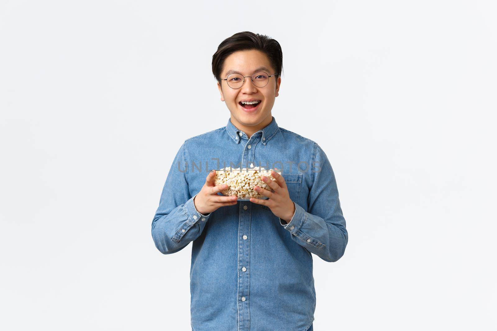 Leisure, lifestyle and people concept. Smiling cute asian man prepared bowl of popcorn for movie night, standing upbeat and grinning, watching TV premier, standing white background by Benzoix