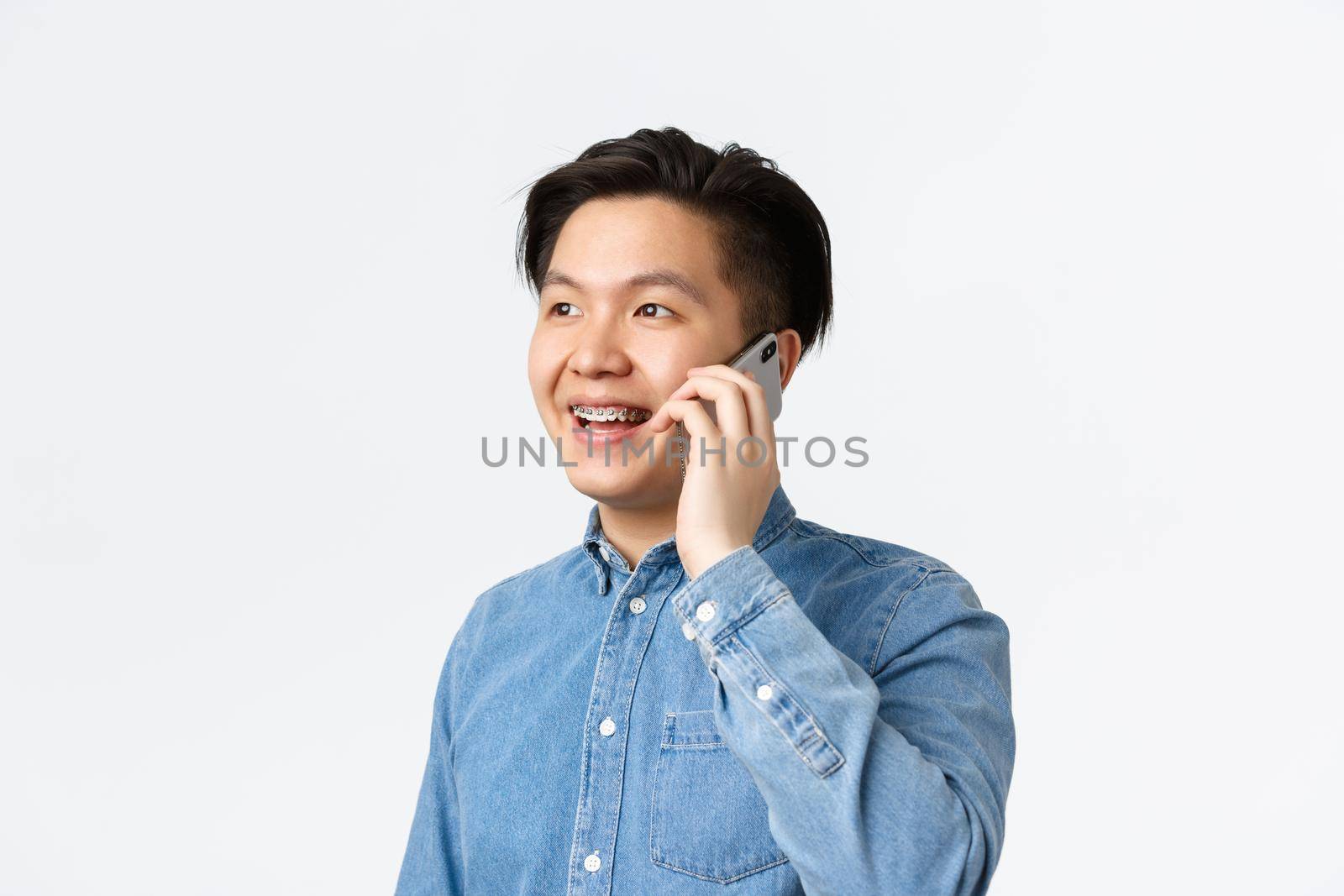 Close-up of happy friendly-looking asian man with braces, smiling and looking away while talking on phone with happy expression, standing white background have conversation.