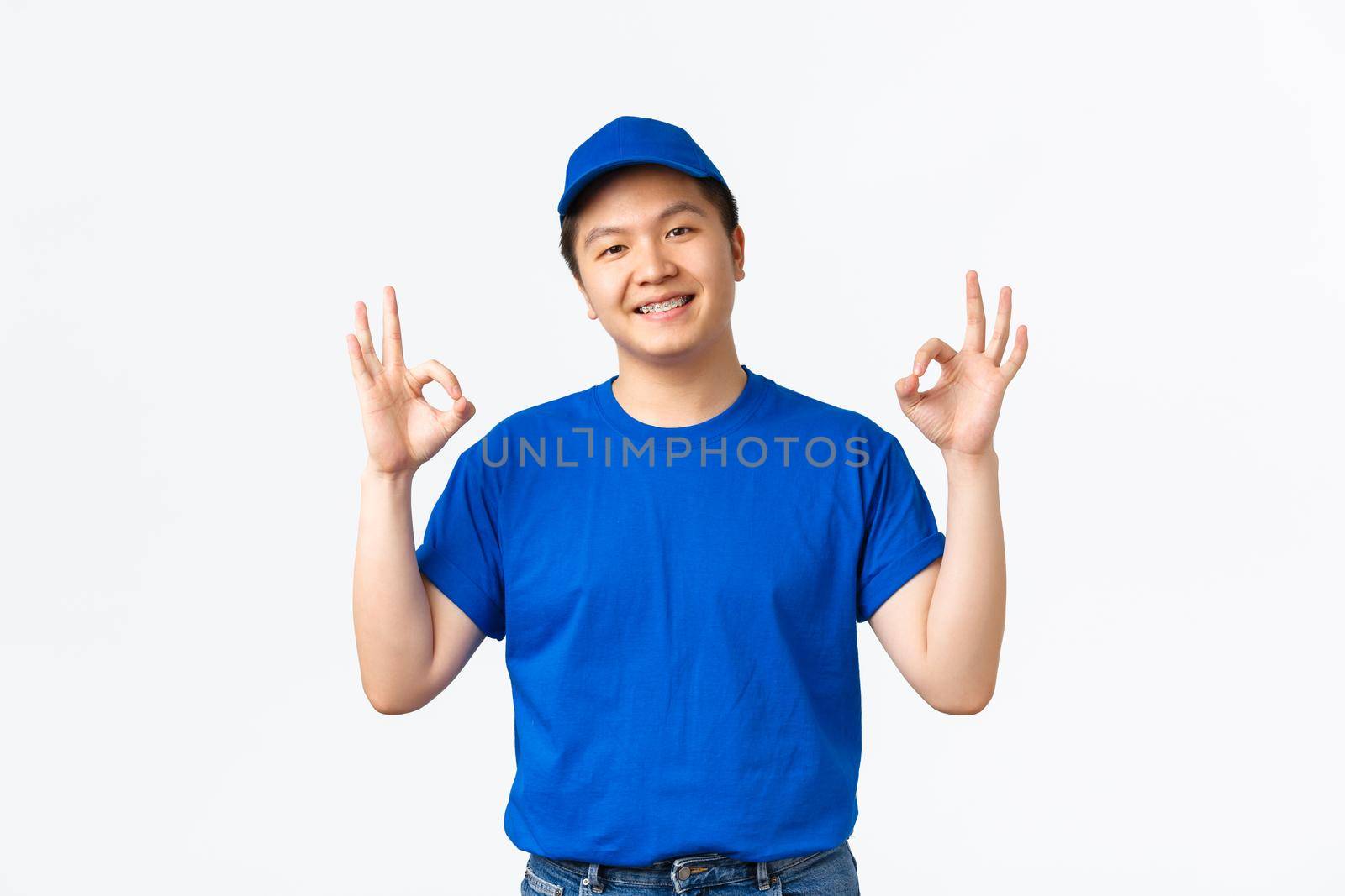 Smiling confident asian courier in blue uniform, wink and showing okay gesture. Delivery guy say OK, guarantee safety and fast shipping of parcels, man working in mail ensure client.