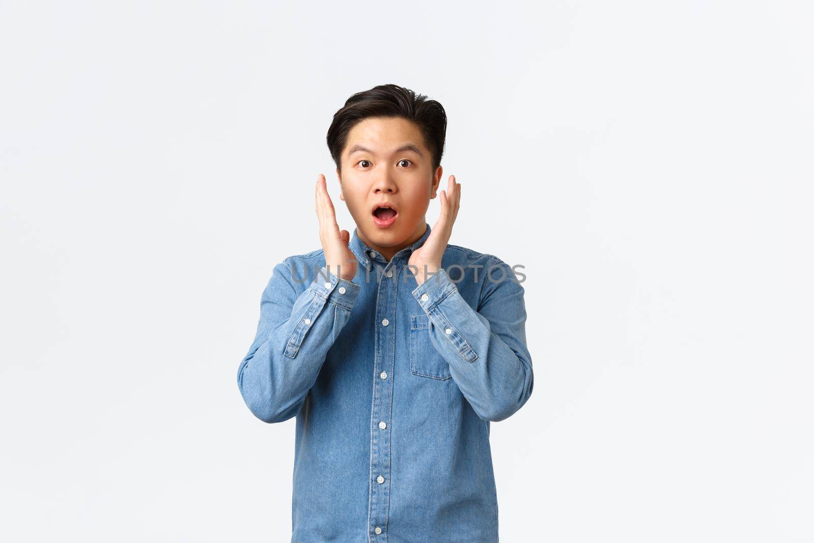 Surprised and impressed asian guy react to big announcement, holding hands near face and gasping, stare at something unbelievable, standing astounded over white background.