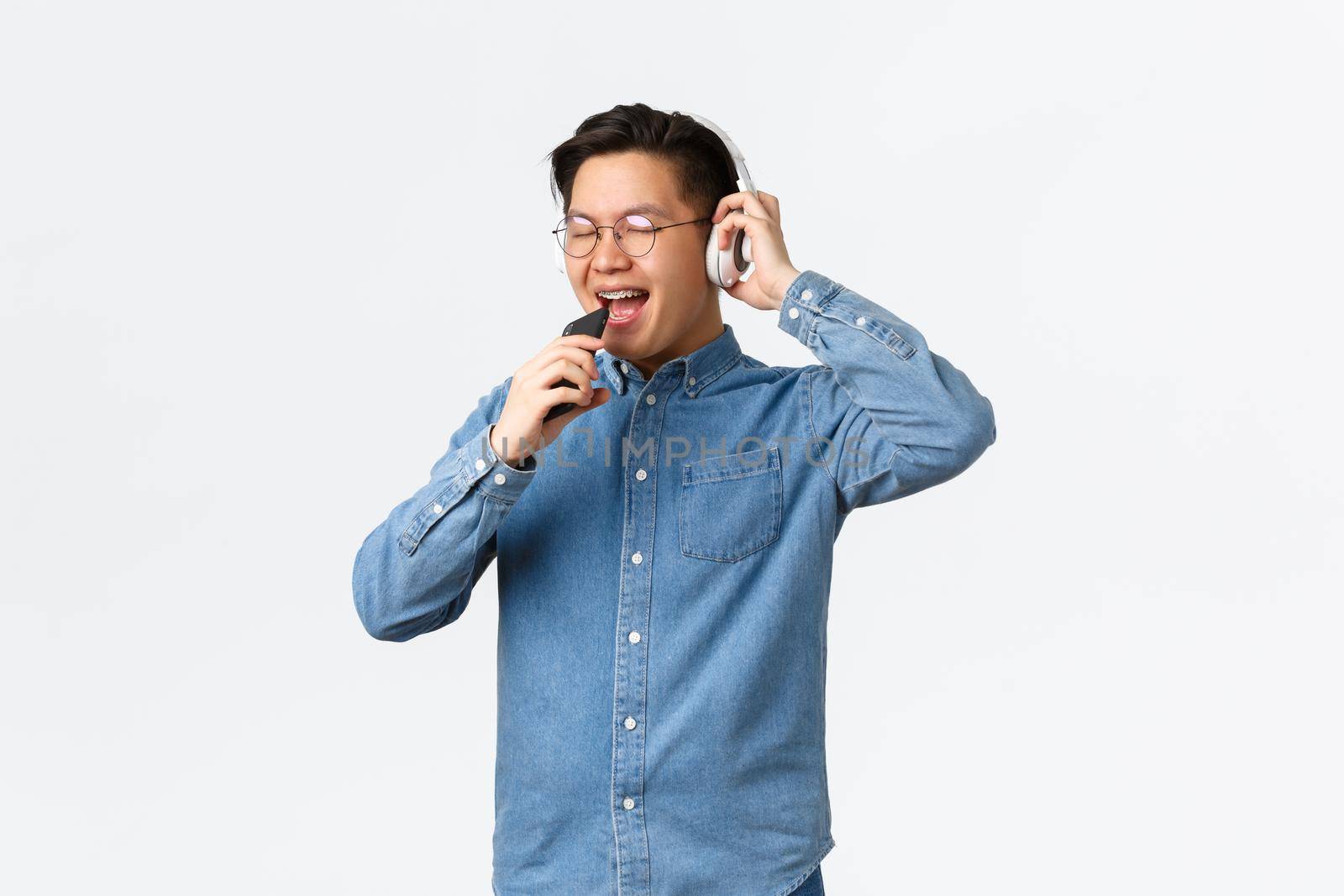 Lifestyle, leisure and technology concept. Carefree happy asian young man playing karaoke app, using wireless headphones, singing in mobile phone dynamic, standing white background.