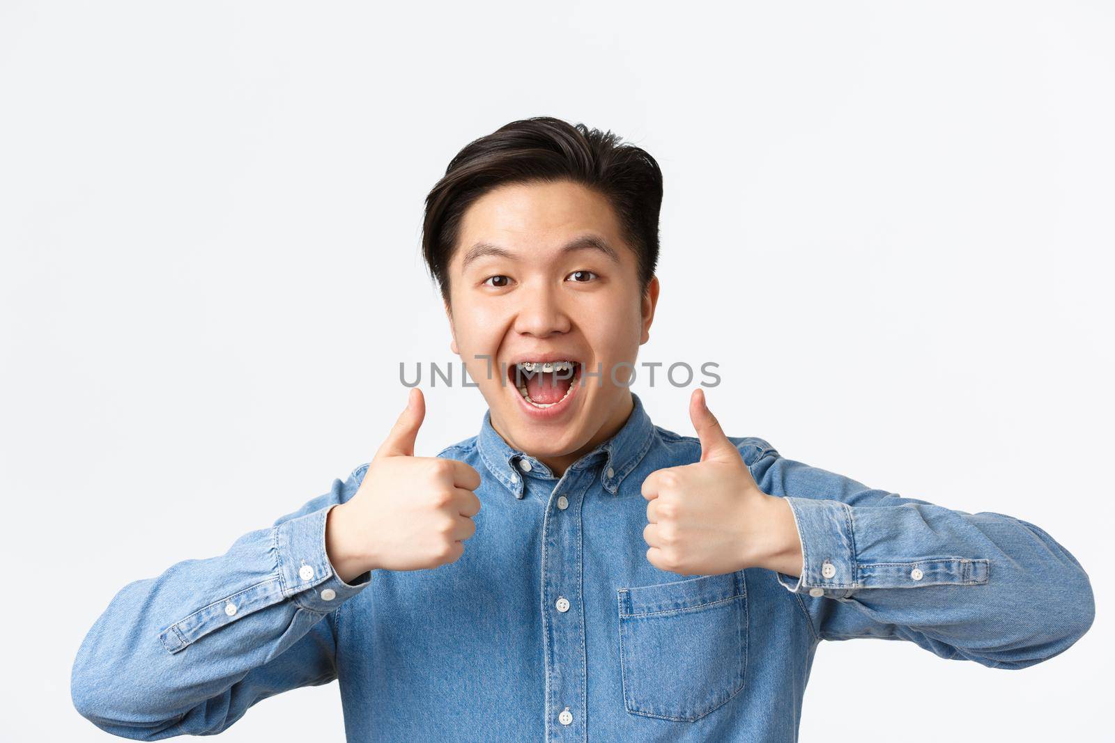 Close-up of successful winning, happy asian man with teeth braces smiling broadly and showing thumbs-up in approval, praising great work, saying well done, standing white background pleased.
