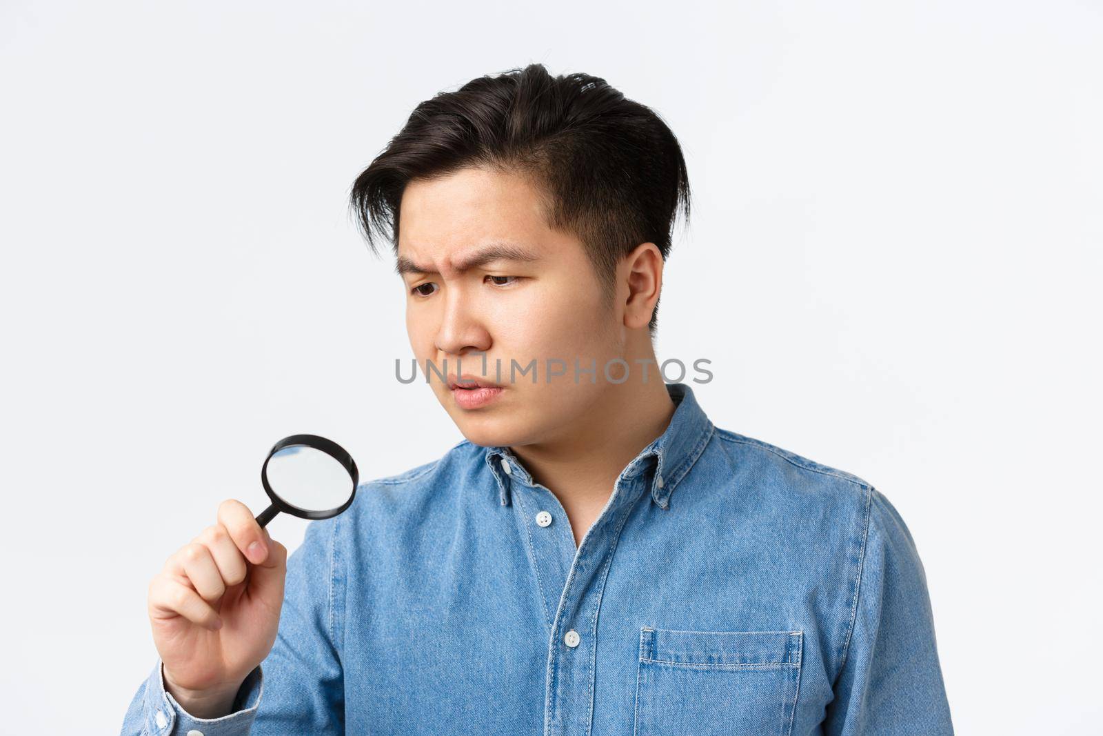 Close-up of serious-looking focused asian man studying something, searching with magnifying glass, looking through it down with concerned face, standing white background.