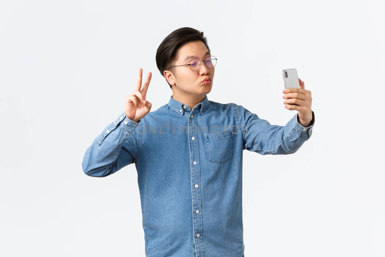 Cute and funny asian young guy pouting silly, taking selfie on smartphone, using photo filter app to change appearance, shooting himself with peace sign and kiss, white background.