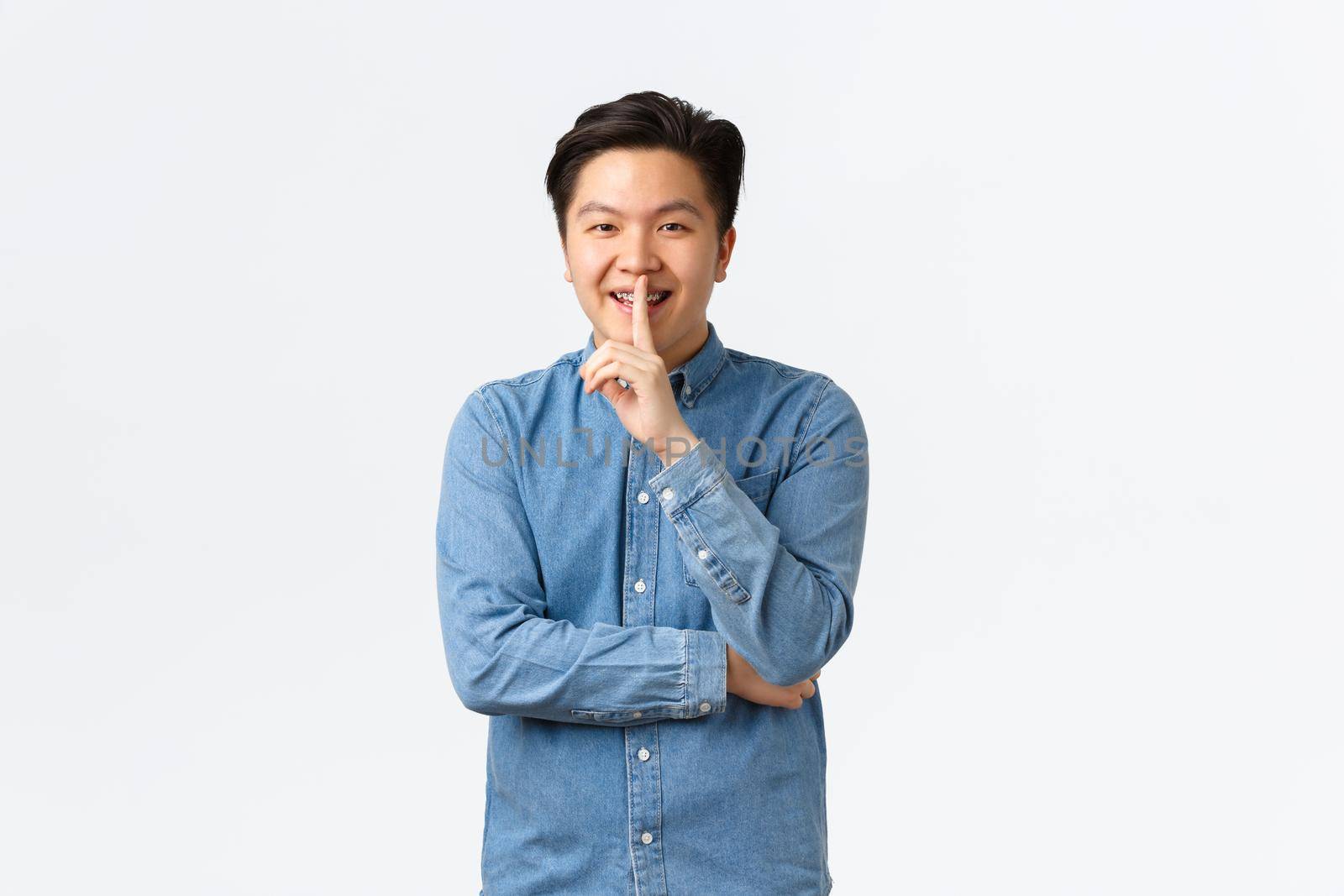Smiling cunning asian man preparing surprise, asking keep quiet, shushing or hushing at person, press index finger to lips, promise not tell, gossiping over white background, whispering.