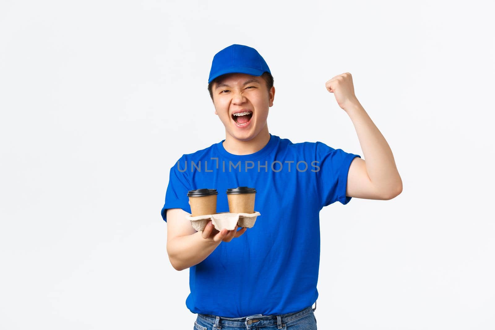 Online orders, takeaway and employees concept. Triumphing cheerful asian male courier in blue uniform bring coffee to client home without spill, rejoicing, shouting hooray or yes, white background.
