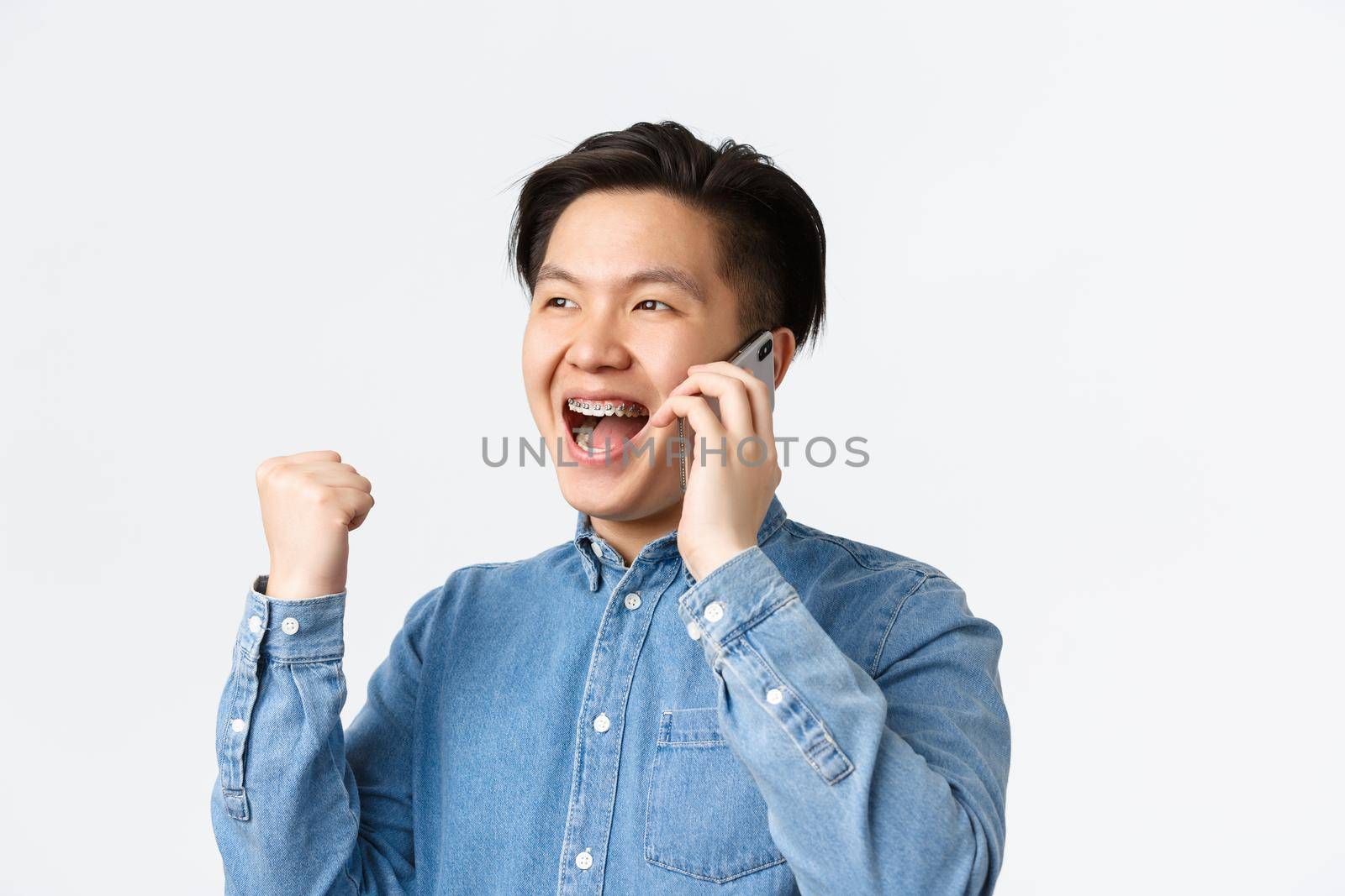 Close-up of winning successful asian businessman in shirt, rejoicing while having conversation over mobile phone, fist pump and smiling, receive good news, standing white background by Benzoix