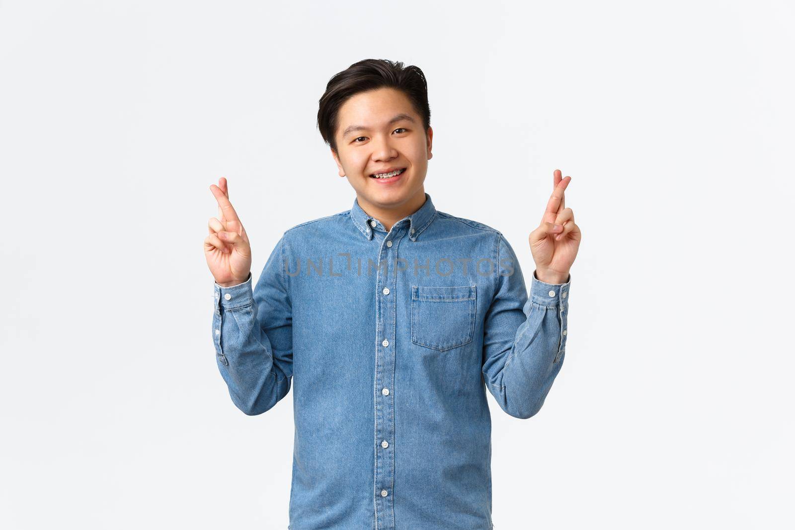 Smiling cute asian man having faith, believe dreams come true, cross fingers good luck, praying or making wish, standing lucky over white background, anticipating positive results by Benzoix