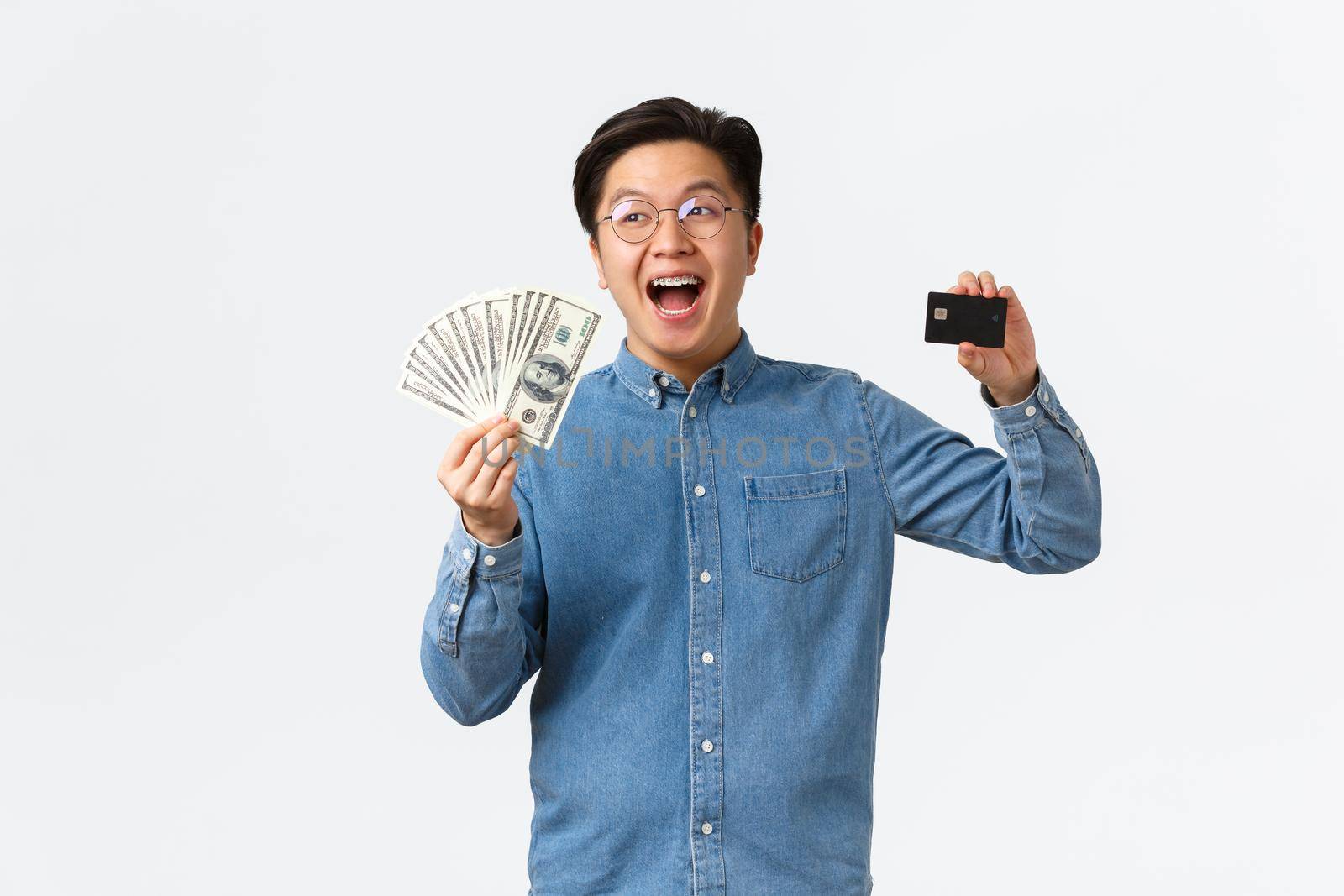 Happy smiling asian man with braces and glasses, laughing cheerfully and showing credit card, holding money, buying something, place cash on bank account, standing white background by Benzoix