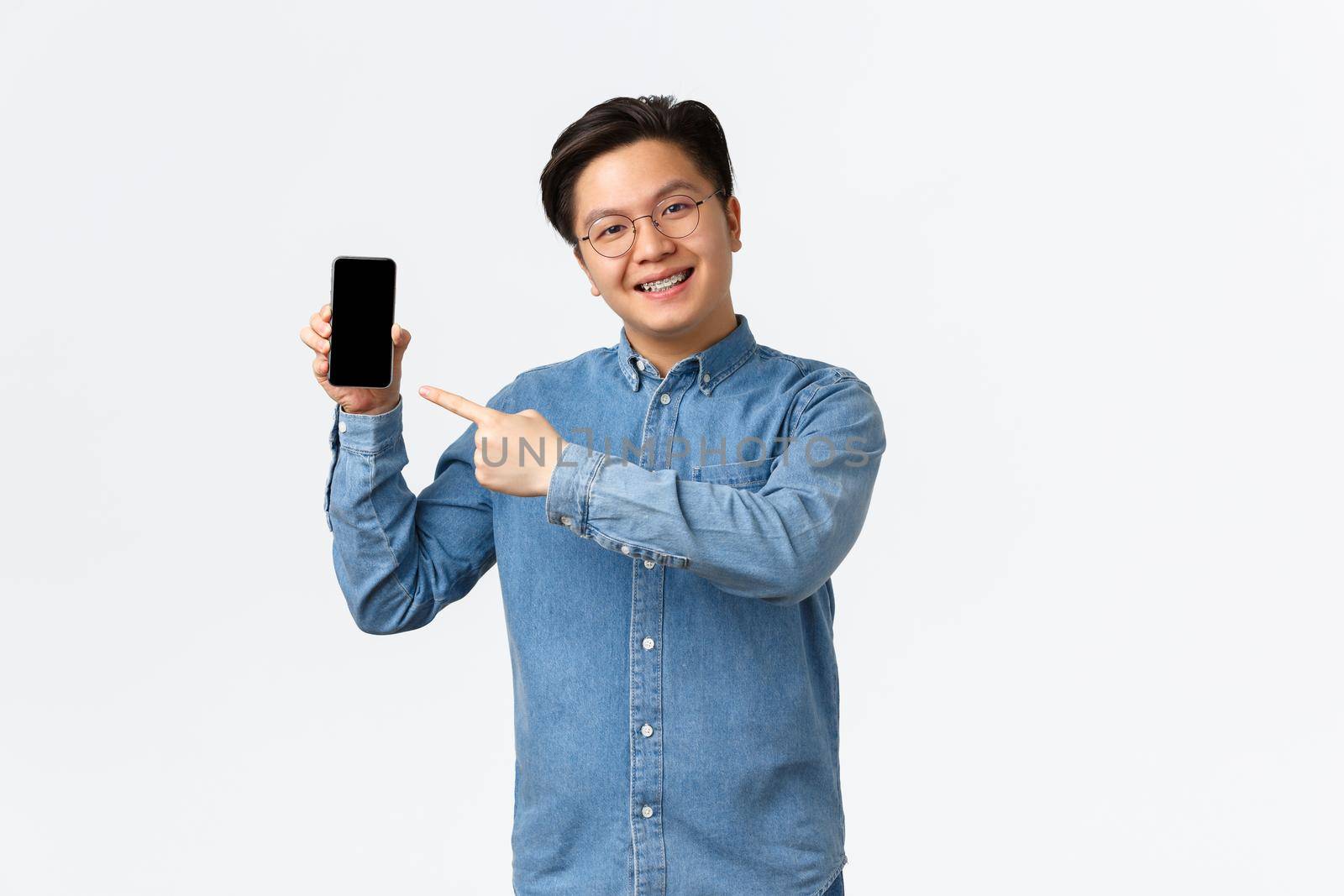 Smiling handsome asian guy with braces and glasses, pointing finger at smartphone screen. Man showing promo or application on mobile phone display, promote site, standing white background.