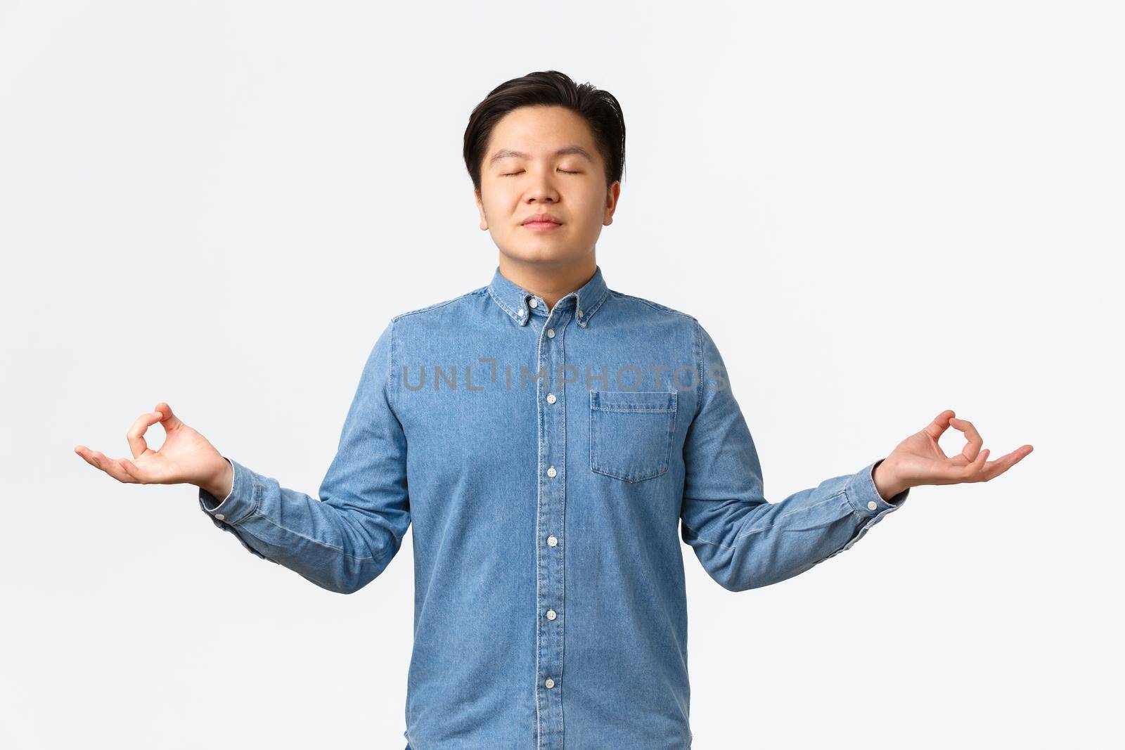 Relieved and relaxed smiling asian man with closed eyes meditating, feeling peaceful and happy, release stress, standing over white background with hands spread sideways in nirvana pose.