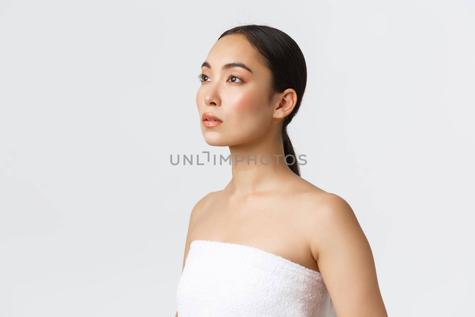 Feminine beautiful asian woman in white towel looking left dreamy. Girl with clean perfect skin, advertising skincare products, bathroom cosmetics, standing white background.