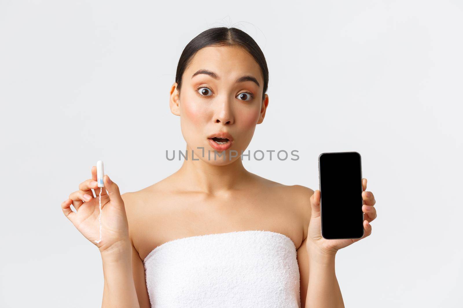 Beauty, personal and intimate care, mobile application concept. Surprised and fascinated asian girl in towel showing tampon and smartphone app for menstrual flow, cycle tracker, white background.