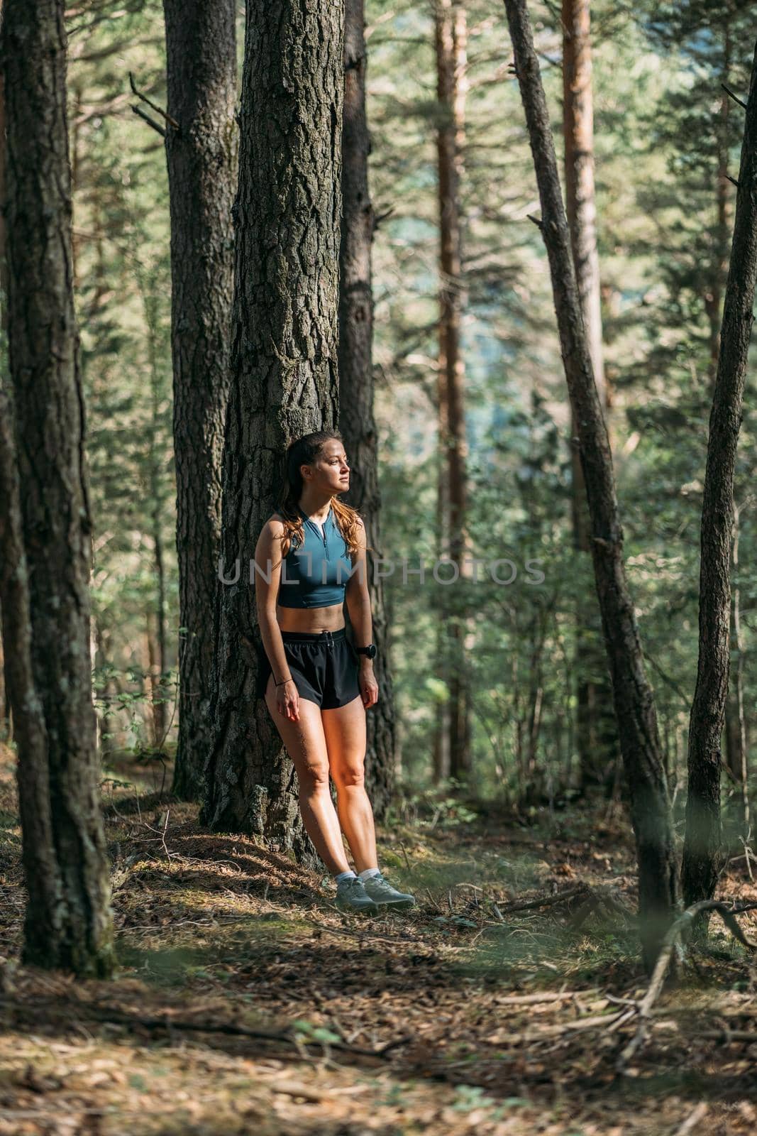 Tired young woman in sportswear leaned on a tree and resting in the forest after working out. Trail running and active life concept. by apavlin