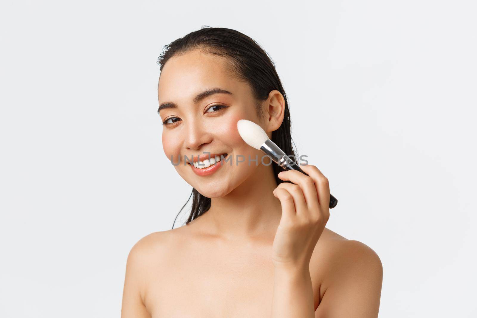 Skincare, women beauty, hygiene and personal care concept. Close-up of gorgeous young asian female using makeup brush on cheeks and smiling, standing naked over white background.