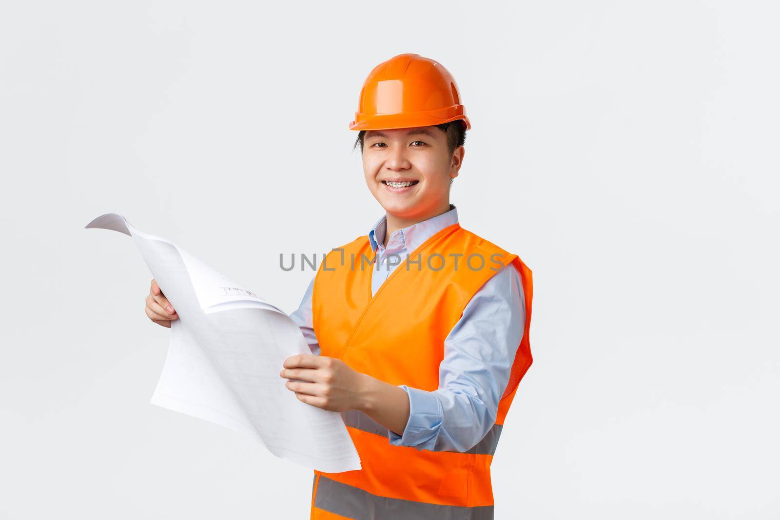 Building sector and industrial workers concept. Confident smiling asian architect, chief engineer in helmet and reflective jacket holding blueprints, inspecting enterprise, white background.