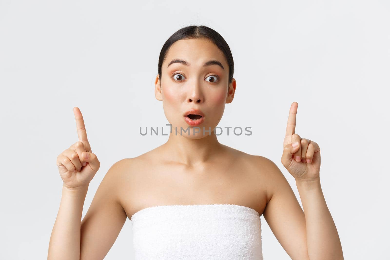 Beauty, cosmetology and spa salon concept. Excited gorgeous asian girl in white towel around naked body pointing fingers up, say wow in amazement as see awesome discounts by Benzoix