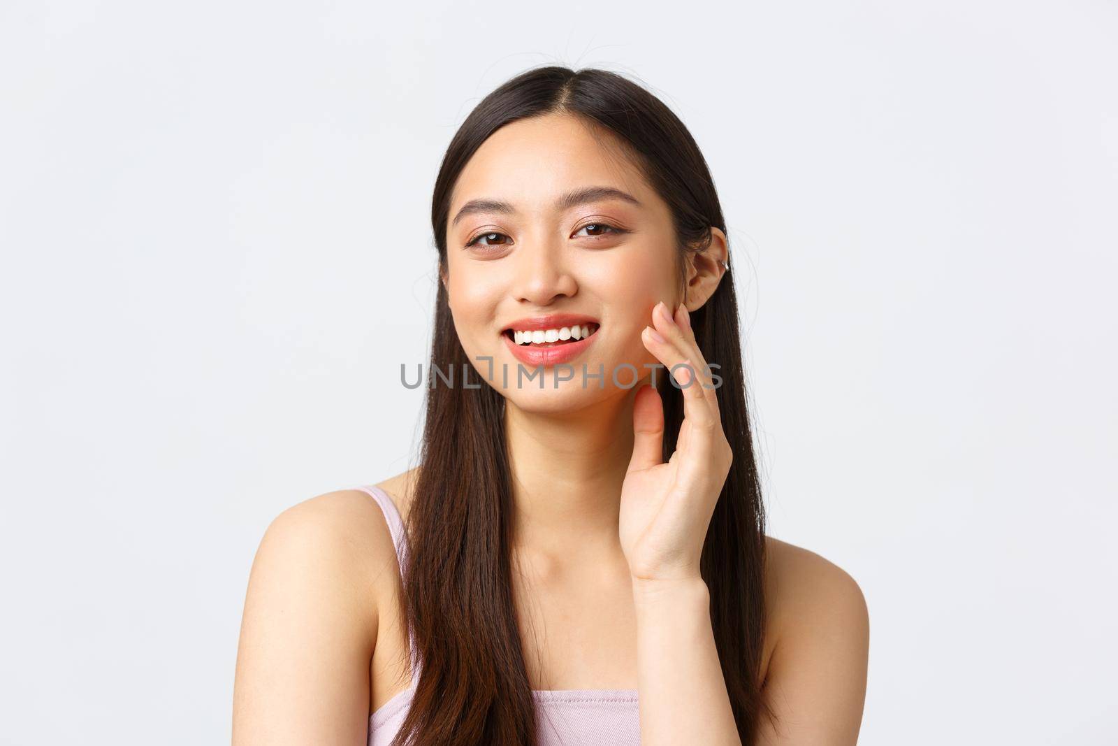Beauty, fashion and people emotions concept. Beautiful asian girl trying new skincare routine product or makeup, smiling happy and satisfied as touching skin gently, white background.