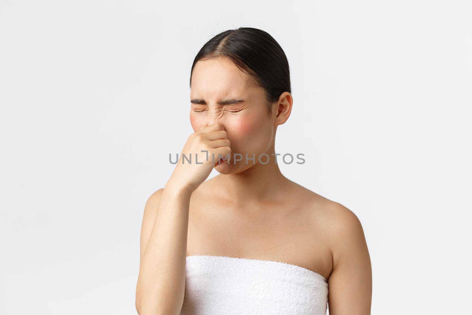 Beauty, cosmetology and spa salon concept. Close-up of young asian woman in bath towel feeling cold after shower, sneezing and cover nose with hand, standing white background.