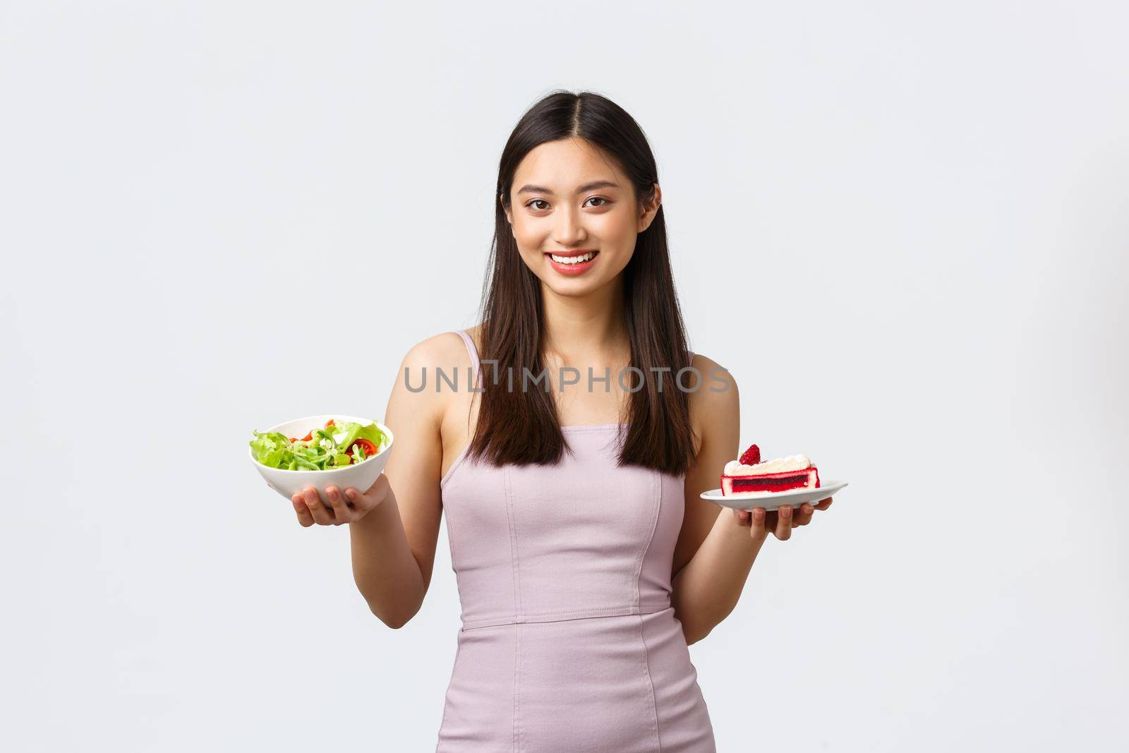 Healthy lifestyle, leisure and food concept. Cheerful beautiful asian girl in dress showing bowl with salad and piece of cake, smiling as give advice on nutrition diet, standing white background.