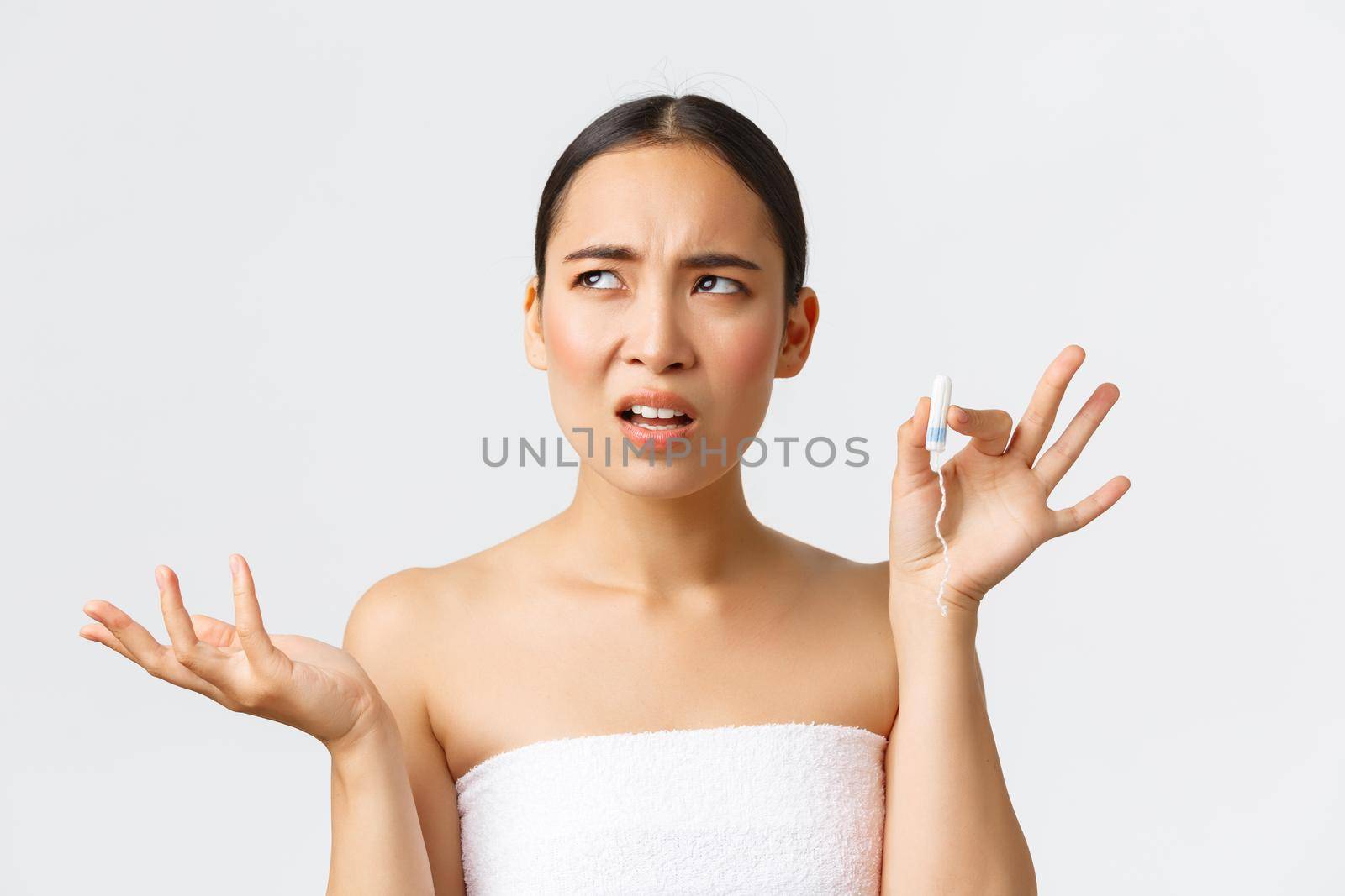 Beauty, personal and intimate care menstrual hygiene concept. Confused and displeased asian woman in bath towel complaining on tampons, raising hand and look away, use menstrual cups.