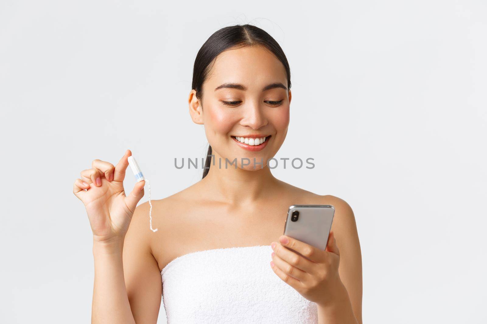 Beauty, personal and intimate care, mobile application concept. Smiling pretty asian girl having period, wear towel, looking at menstrual cycle tracker app on smartphone, holding tampon by Benzoix