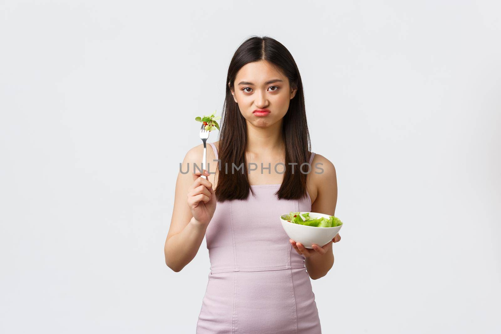Healthy lifestyle, leisure and people emotions concept. Gloomy pouting asian girl dislike eating vegetables, holding bowl with salad, grimacing as unwilling sit on diet, white background.