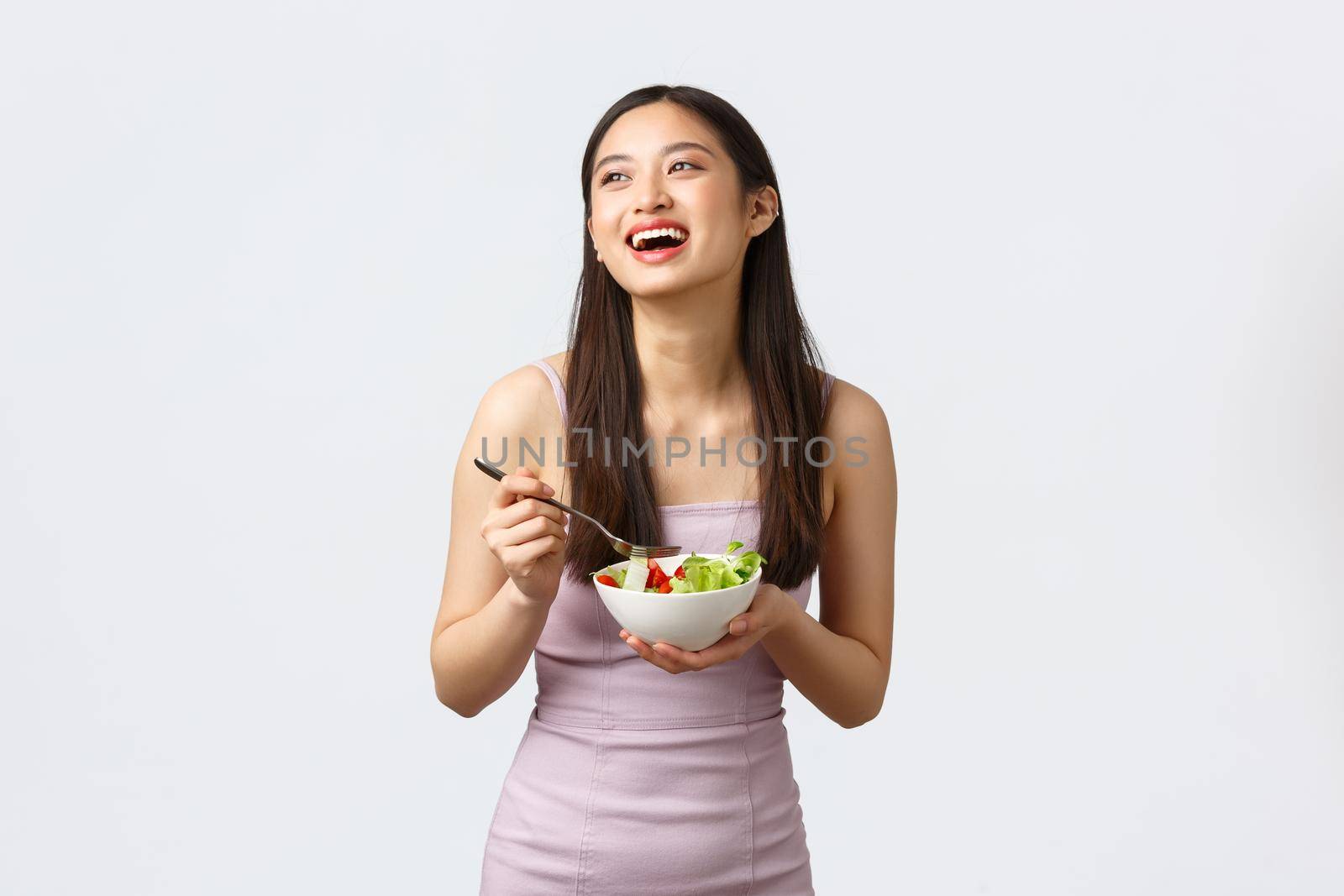 Healthy lifestyle, leisure and people emotions concept. Gorgeous happy asian girl smiling and laughing while eating salad, staying fit for summer, wearing evening dress, white background.