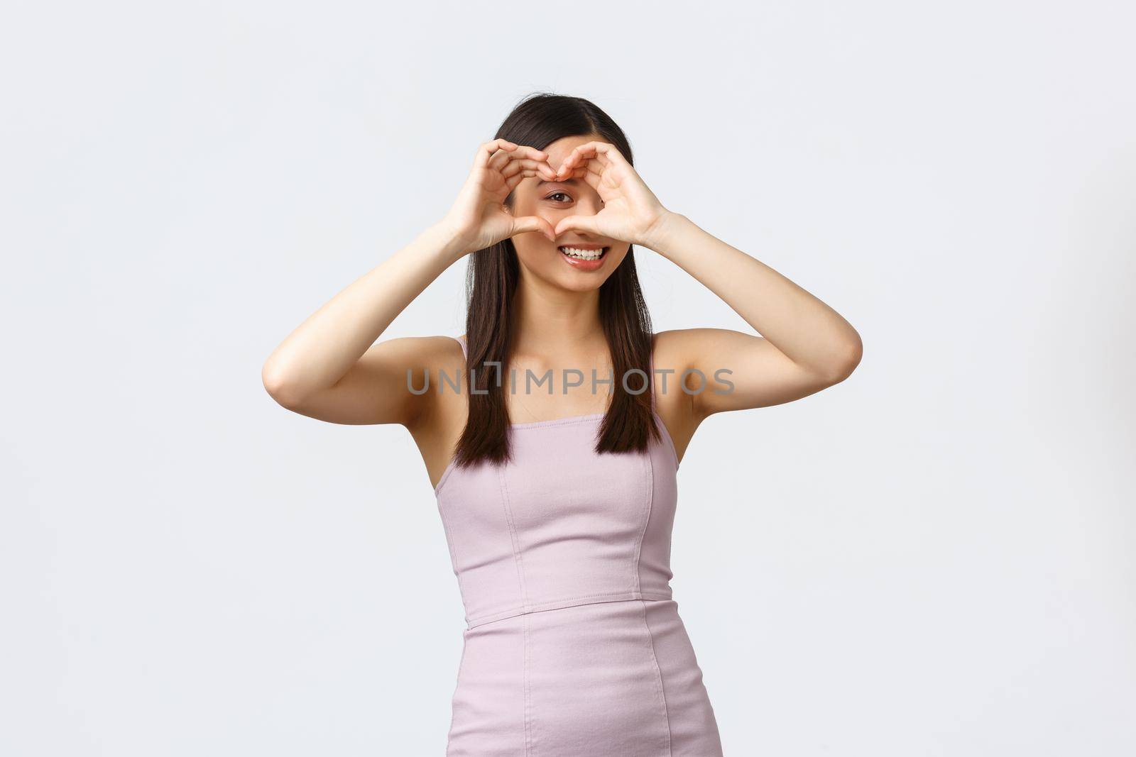 Luxury women, party and holidays concept. Lovely silly asian woman in evening dress, looking through heart sign and smiling, express sympathy or love, feeling romantic, white background by Benzoix