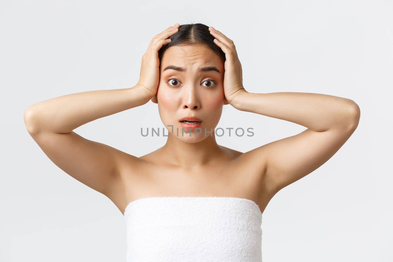 Beauty, cosmetology and spa salon concept. Troubled cute asian woman in panic grab head and looking confused camera. Overwhelmed korean girl in bath towel react to shocking news, stare concerned.