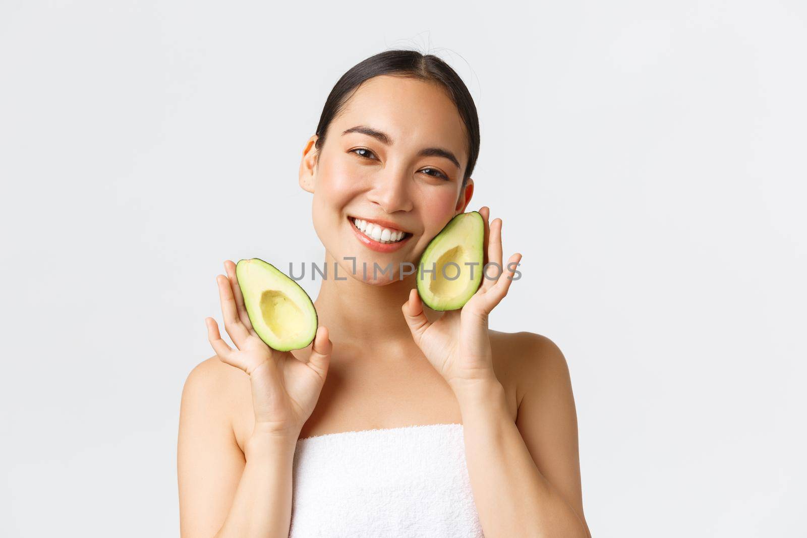 Beauty, personal care, spa and skincare concept. Tender cute asian female in bath towel showing avocado and smiling satisfied, nourish and treat skin with face masks or cream, white background.
