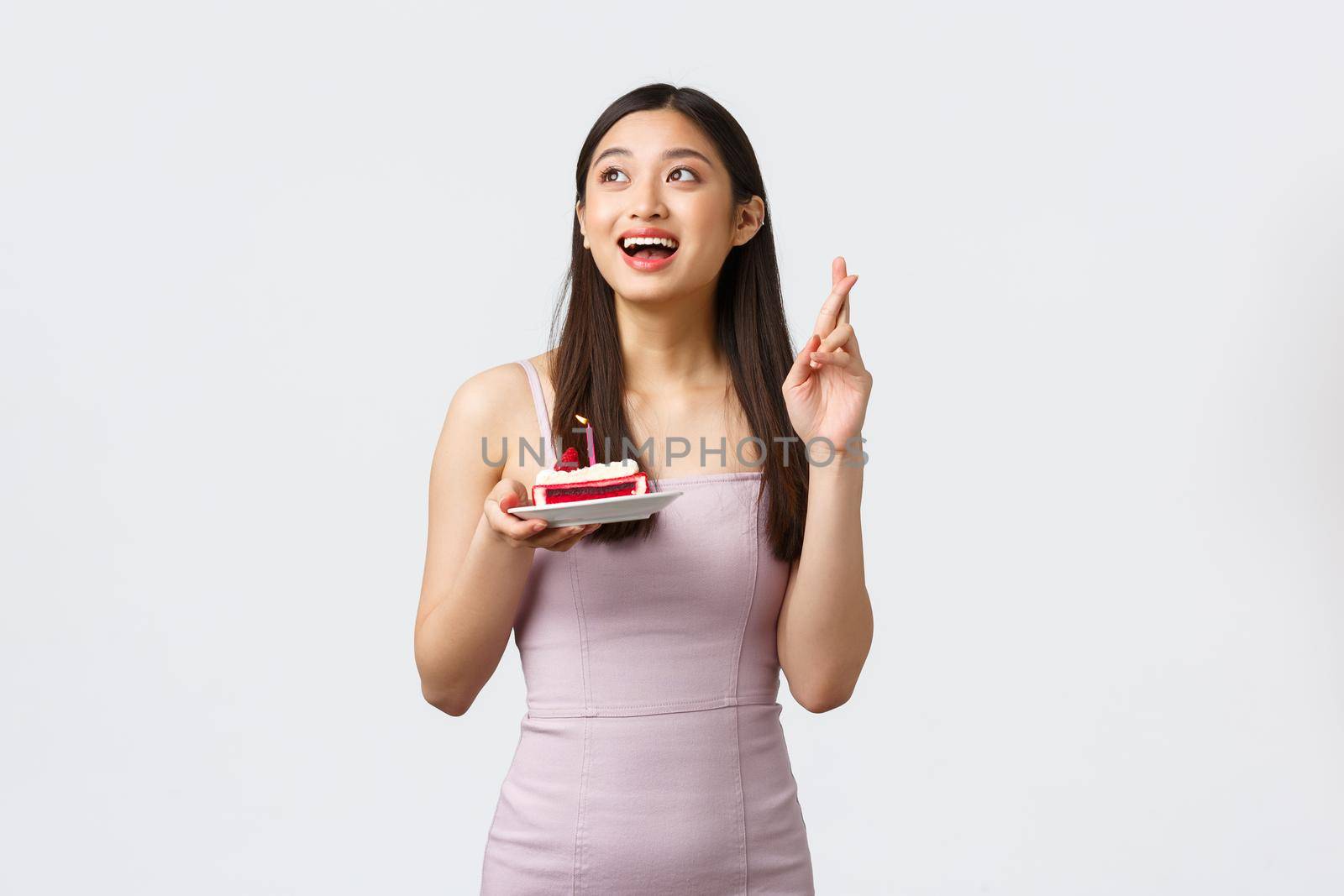 Celebration, party and holidays concept. Hopeful dreamy upbeat birthday girl in dress, cross fingers good luck looking upper left corner, holding b-day cake, white background.