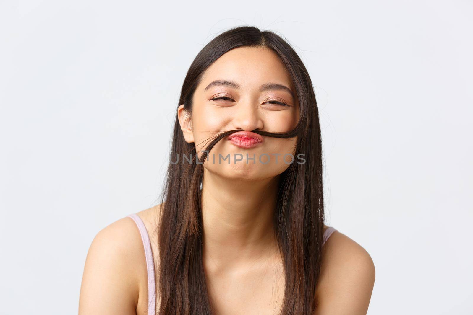 Concept of beauty, fashion and makeup products advertisement. Close-up portrait of happy silly asian girl making fake moustache out of hair strand, smiling cheerful, white background.