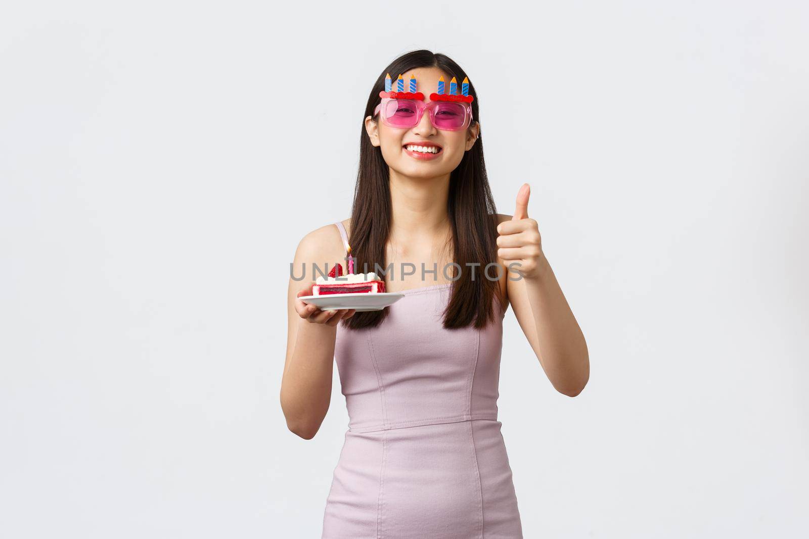 Celebration, party and holidays concept. Joyful smiling asian woman in glasses and dress, show thumbs-up, celebrating own birthday, making wish on b-day cake, white background.