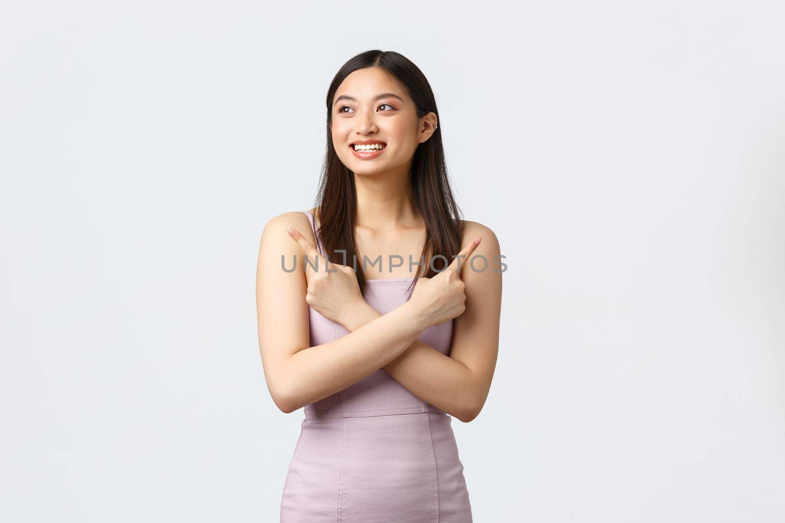 Luxury women, party and holidays concept. Carefree smiling, attractive asian woman in stylish dress, pointing fingers sideways and looking pleased left as made her choice, white background.