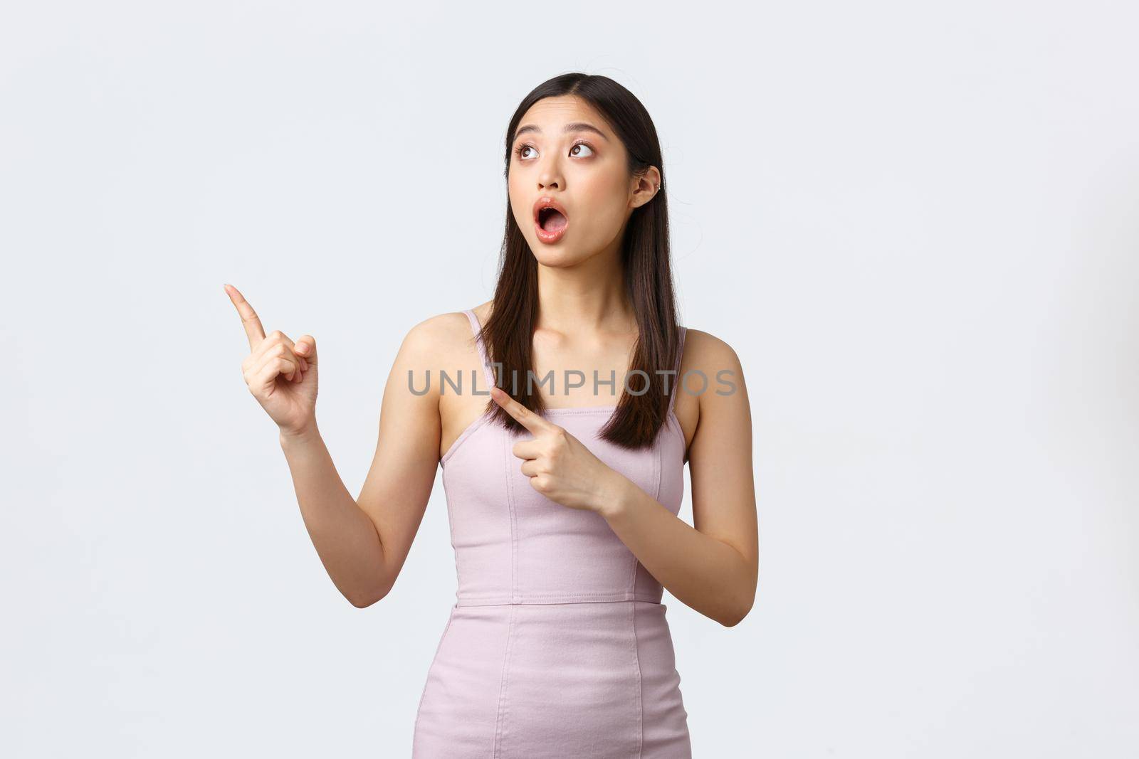 Luxury women, party and holidays concept. Impressed and shocked beautiful asian woman in evening dress, drop jaw and pointing upper left corner with astonished expression, white background.