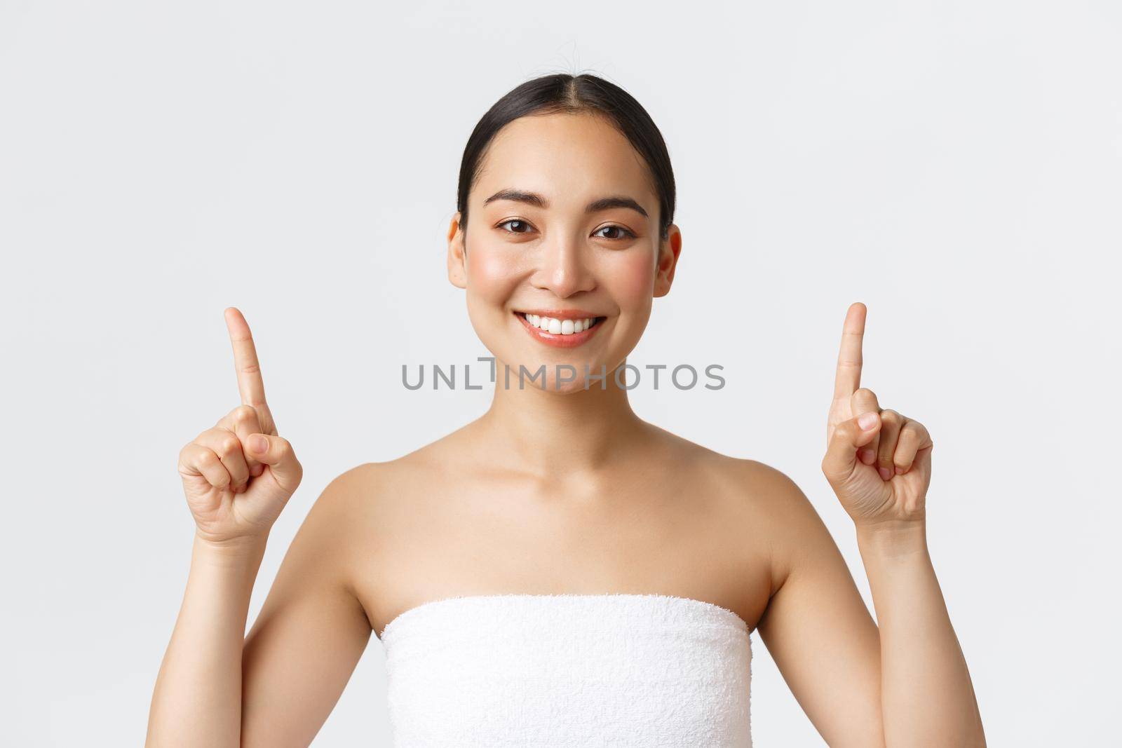 Beauty, cosmetology and spa salon concept. Happy pleased asian female in towel smiling satisfied and pointing fingers up, recommend massage therapy, skincare products, white background by Benzoix