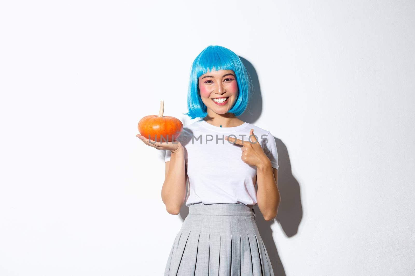 Cute smiling asian woman in blue wig pointing finger at small pumpkin, celebrating halloween, standing over white background.