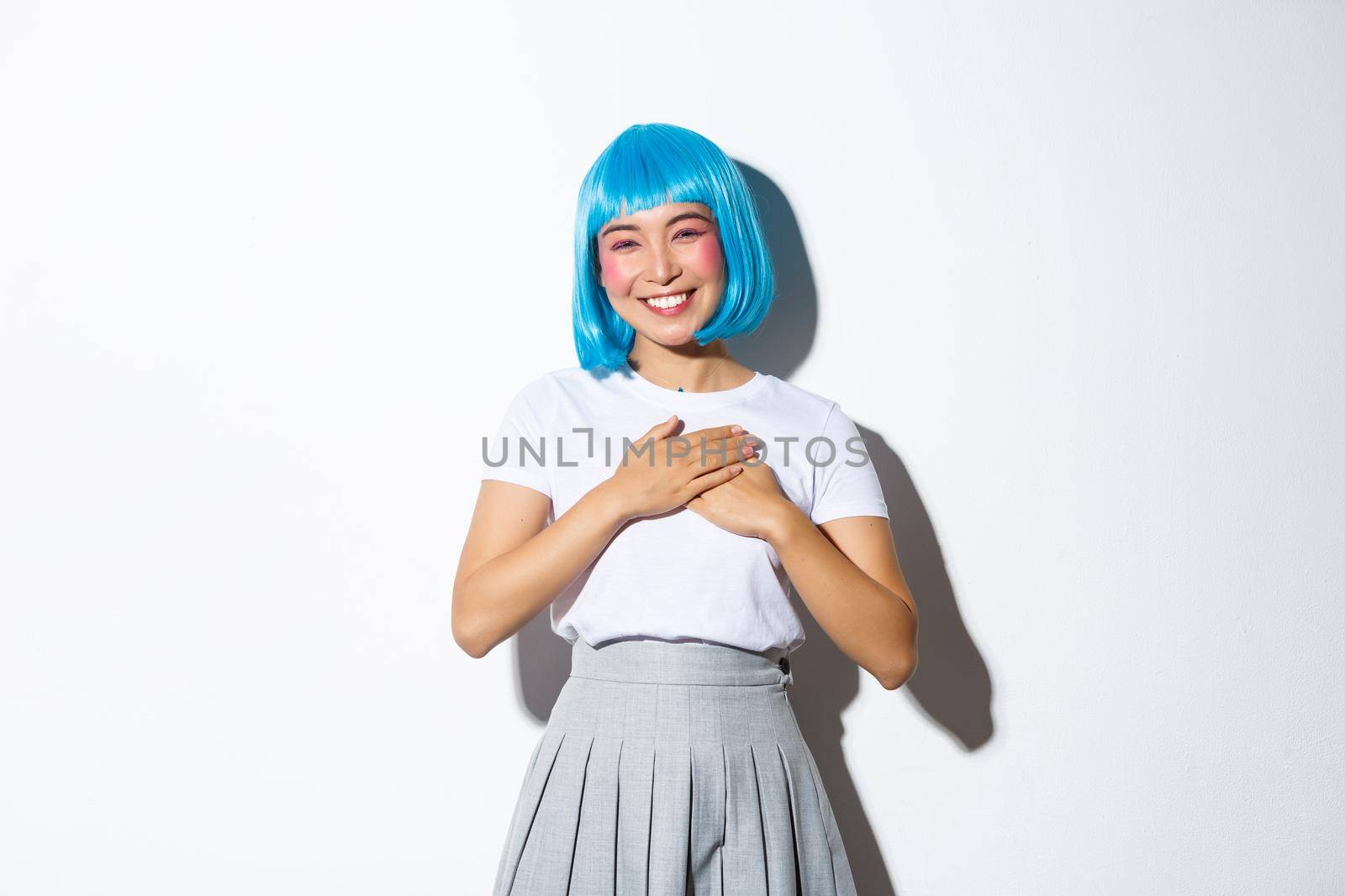 Image of thankful smiling asian girl in halloween costume and blue party wig, holding hands on heart and looking grateful, thanking for something, standing over white background.