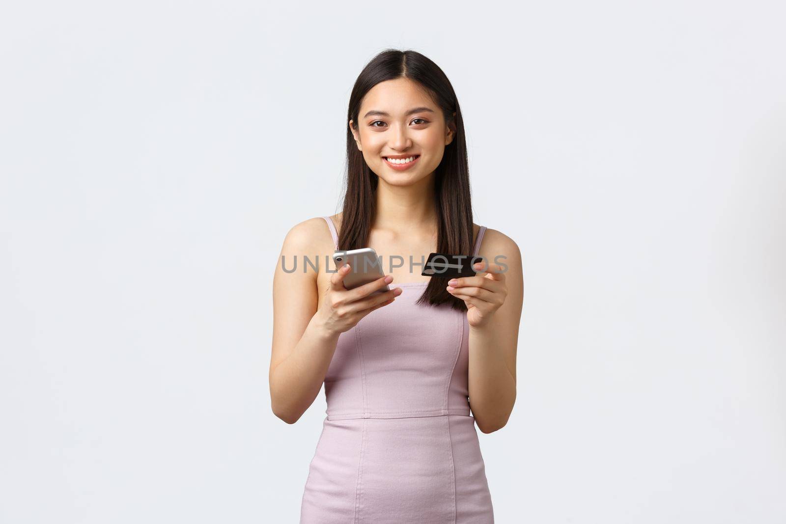 Luxury women, party and holidays concept. Smiling attractive asian girl in evening dress, holding credit card and smartphone, looking camera as making order in shopping app, contactless payment by Benzoix