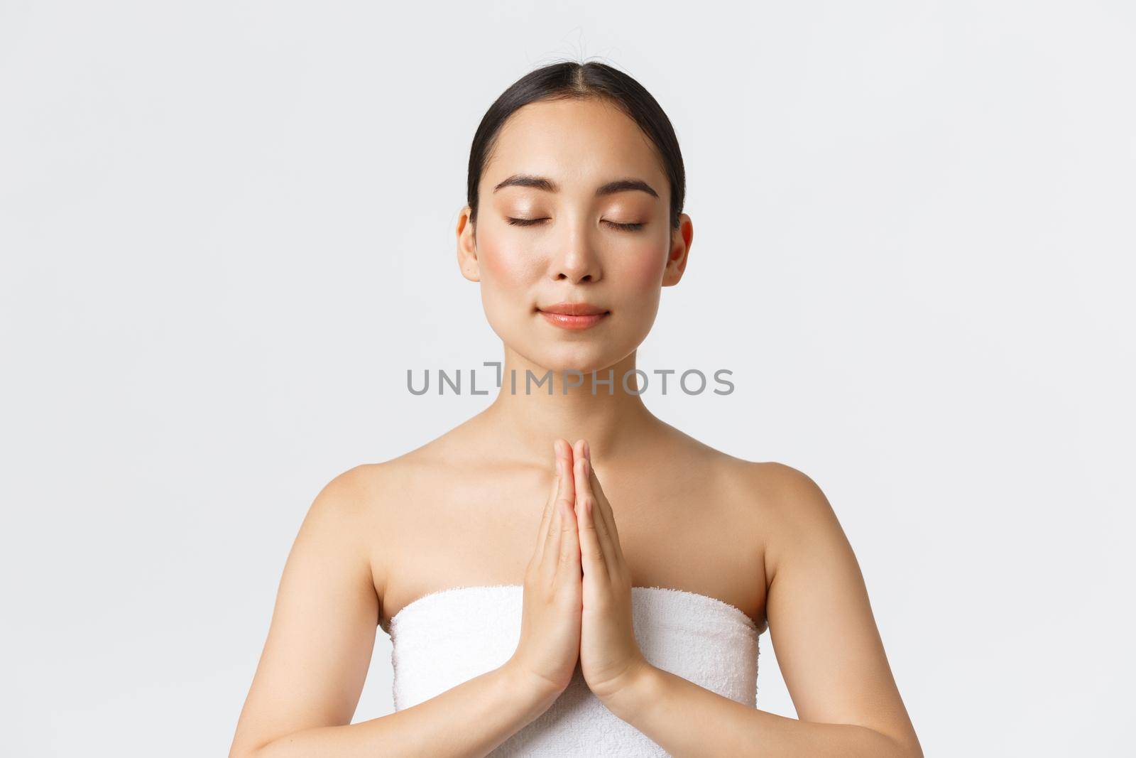 Beauty, cosmetology and spa salon concept. Relaxed sensual pretty asian girl in white towel press hands together and close eyes as meditating, namaste pose while enjoying beauty procedures.