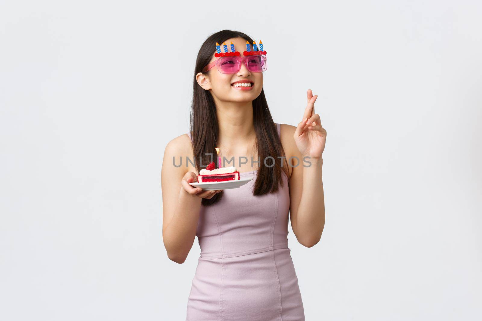 Celebration, party and holidays concept. Beautiful dreamy asian girl in funny sunglasses, cross fingers over b-day cake, making wish, smiling happy and looking upper right corner.