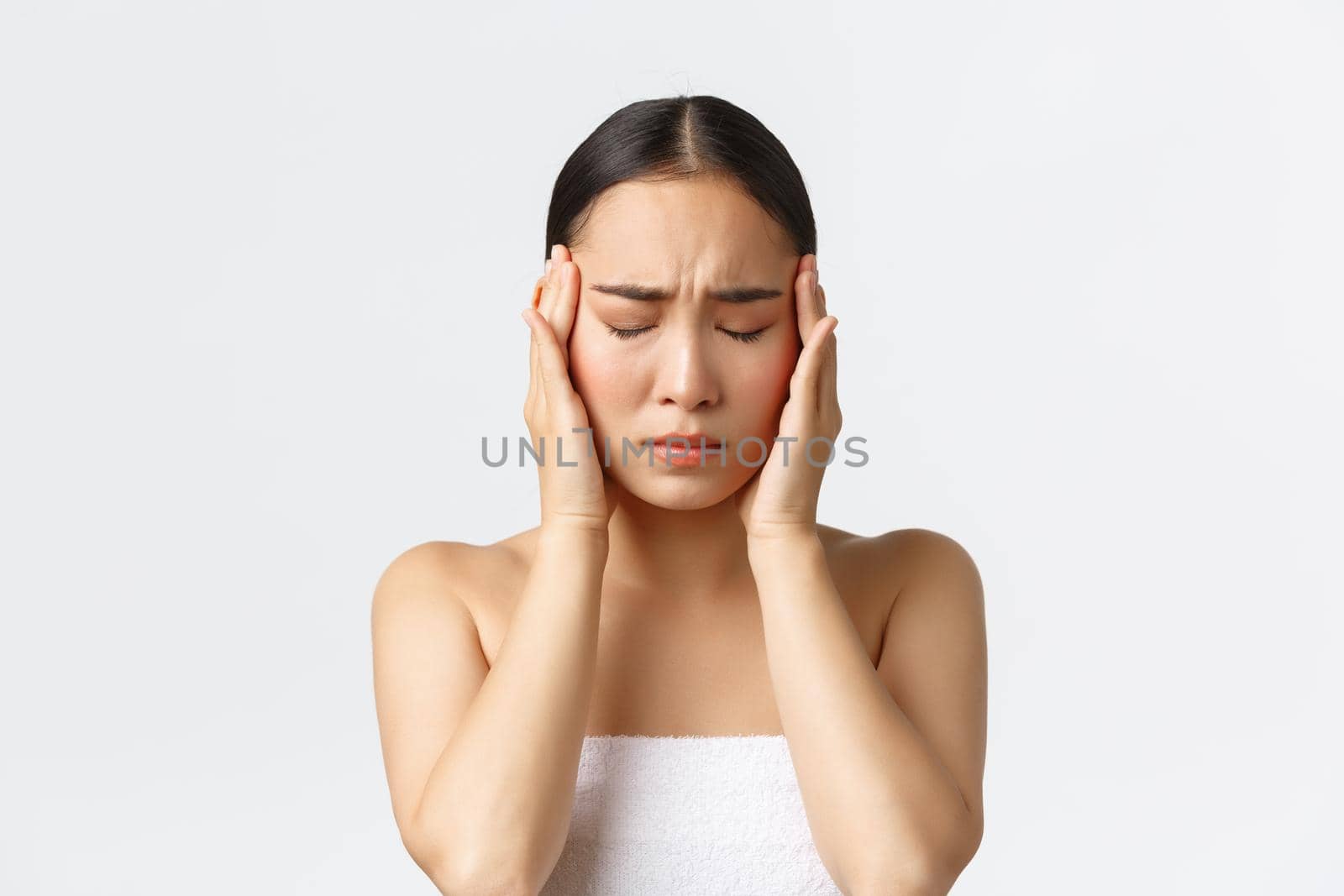 Beauty, cosmetology and spa salon concept. Young uneasy asian woman in bath towel feeling migraine, touching temples and grimacing from painful feeling in head, suffer headache, white background.